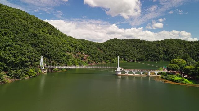 Panning left across white Haneuldari pedestrian bridge at Chopyeong lake in Jincheon, where arched causeway and floating walkways join Chorong gil shoreline trail below forested hills