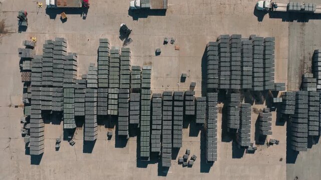 Top-down drone perspective of neatly stacked cinder blocks and flatbed delivery trucks parked inside a large commercial manufacturing yard.