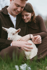 Obraz premium Cheerful couple outdoors with a white goat kid, warm clothes, genuine smiles, sunlit grass meadow, relaxed pose, candid lifestyle moment, natural colors, rustic vibe, playful affection