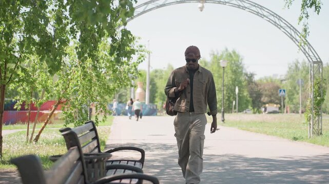 Man walks along tree lined path in urban park carrying bag and adjusting his clothing. Man approaches camera while other people walk ahead on same pathway. scene features green foliage