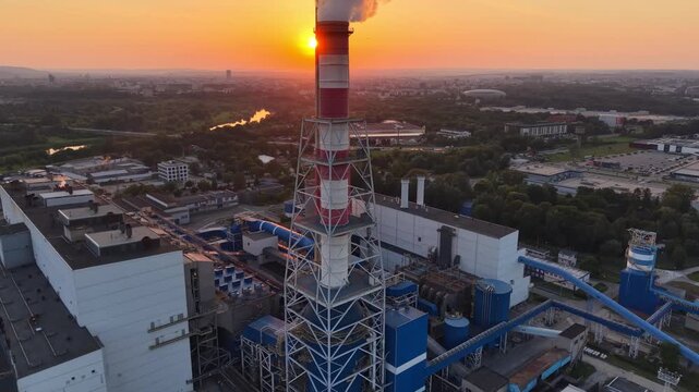 Aerial view of thermal power plant at sunset, showcasing smokestack emitting steam, surrounding landscape, and industrial structures with vibrant sky colors and sun setting in the background