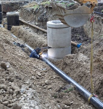 Industrial welder joining underground pipeline near concrete manholes in deep trench
