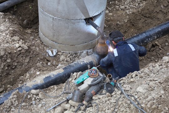 Professional welder joining steel pipes in excavation trench at utility construction site
