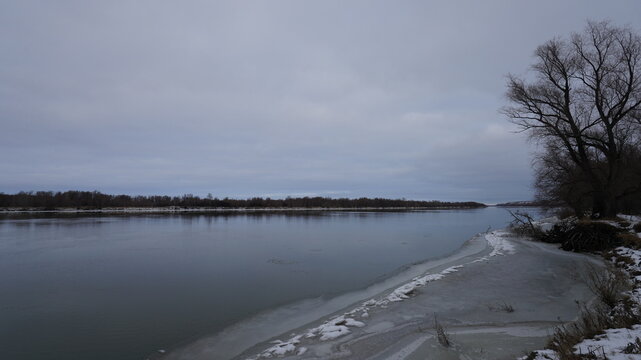 Wide river in late autumn or early winter under a cloudy sky, first ice and snow on the riverbank