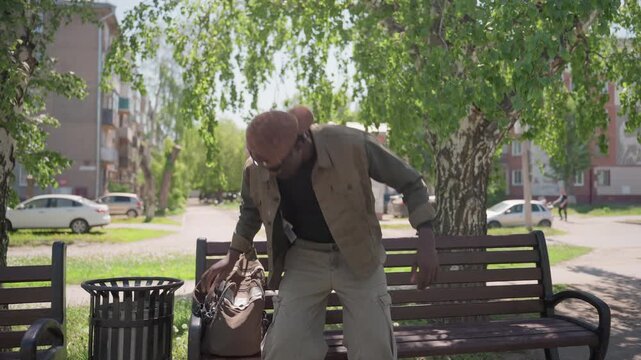 Man with backpack approaches and sits on wooden bench in urban park adjusting his bag before relaxing. scene features green trees overhead parked cars along street behind him apartment