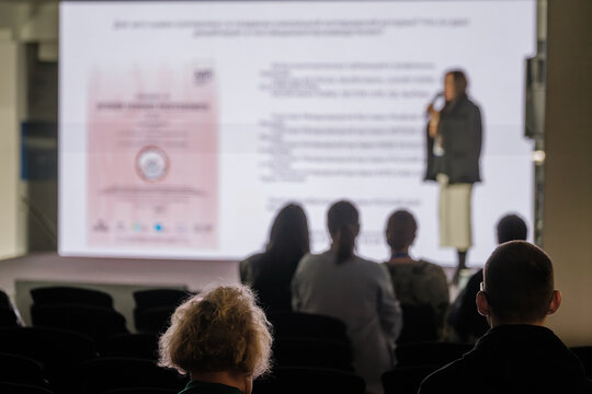 Dark audience silhouettes contrast with projected slides and a standing speaker holding a microphone.
