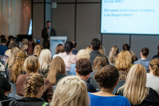 Seated listeners face a male speaker holding a microphone beside projected slides.