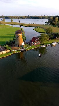 Vertical aerial drone clip of traditional Dutch windmill, river canal, green polder fields, and lake landscape in Warmond near Leiden, South Holland, Netherlands at golden hour sunset