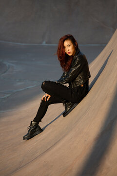 A woman in a leather jacket sits on a concrete ramp outdoors
