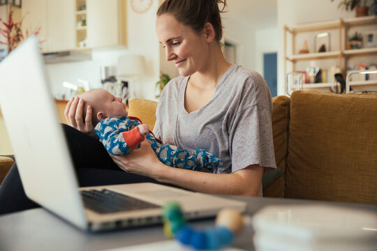 Mother holding her newborn baby at home next to laptop