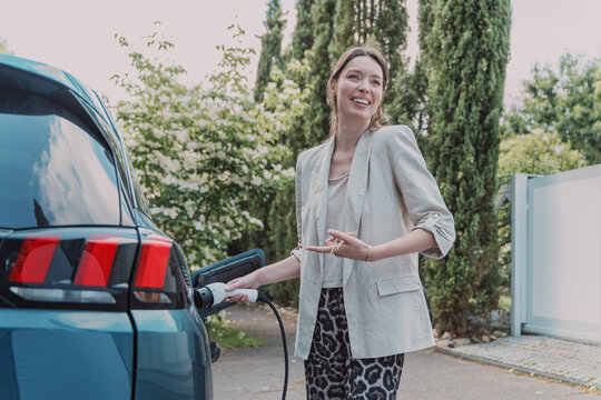Smiling woman is charging her electric vehicle at home outdoors