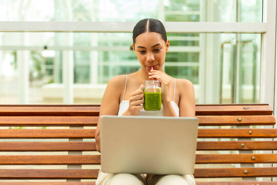 Woman drinks a green smoothie while working on a laptop outdoors