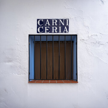 Ventana tradicional con reja y letrero de azulejos de una antigua carnicer&iacute;a en una fachada blanca.