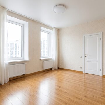 A spacious empty room shows light hardwood flooring, floor to ceiling windows, and sheer curtains overlooking a green outdoor view.