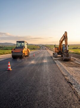 A road roller compacts fresh asphalt at sunset while an excavator works nearby on a highway construction site, creating dust in the golden light.