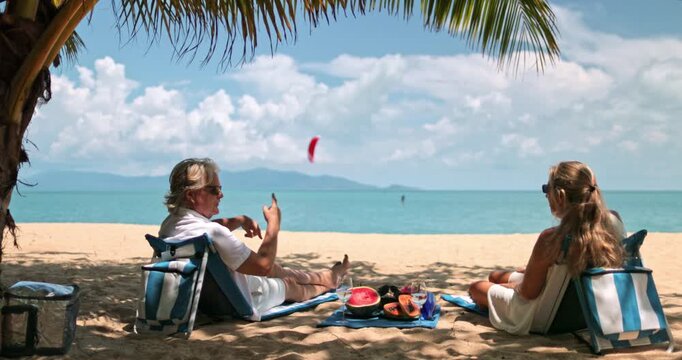 Couple enjoying a relaxing tropical vacation, sitting in beach chairs under a palm tree shadow, having a picnic with fresh fruit by the turquoise ocean. Travel and relax in paradise, Samui, Thailand