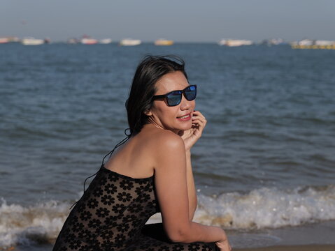 A happy woman wearing sunglasses and a black floral swimsuit enjoys a sunny day at a beach with blue water and boats in the background