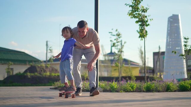 Father teaches little son to ride skateboard in park. Dad supports happy child balancing on board outdoors. Father and son enjoy skateboard practice at sunset. Family bonding on paved path in summer.
