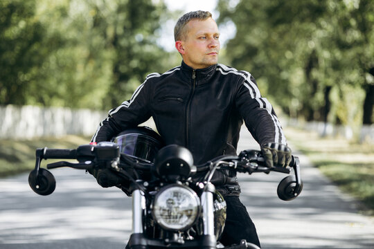 Biker in leather jacket holding a helmet and sitting on a motorcycle, looking to the side on a rural road