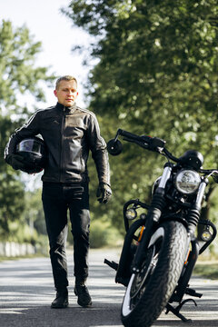 Man in leather jacket and jeans standing on a road, holding a helmet, with his custom motorcycle parked nearby