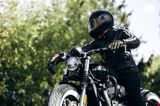 Biker wearing full-face helmet and leather jacket, riding a black vintage custom motorcycle on a road with green trees