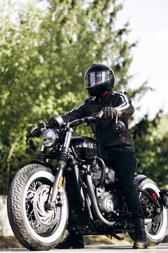 Biker with a black leather jacket and helmet sitting on a classic custom bobber style motorcycle, ready for a ride