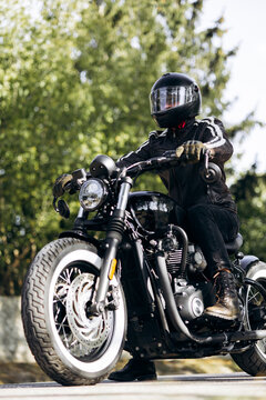Biker wearing a full-face helmet and leather jacket, sitting on a classic black custom motorcycle on an asphalt road