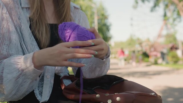 Close up of young woman untangling purple yarn while holding it in her hands wearing light colored lace sleeves and carrying brown bag. scene is outdoors with soft natural lighting