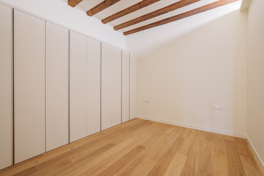 Empty minimalist room with light oak wooden floor, white walls, exposed ceiling beams, and full-height built-in storage cabinets with flush doors, showing warm natural light and clean shadows
