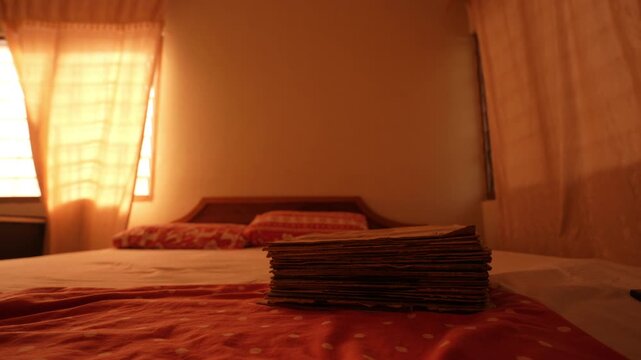 Stack of flattened cardboard record albums rests on a bed in a warmly lit bedroom near curtained windows