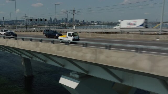 Vehicles cross a Champlain bridge in Montreal over water with a distant urban skyline under daylight, aerial tracking pullback