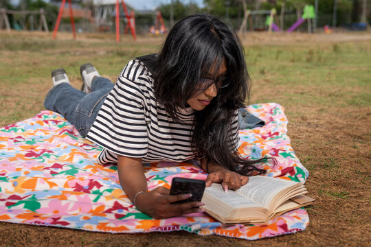 Woman using a smartphone while reading a book on a picnic blanket outdoors