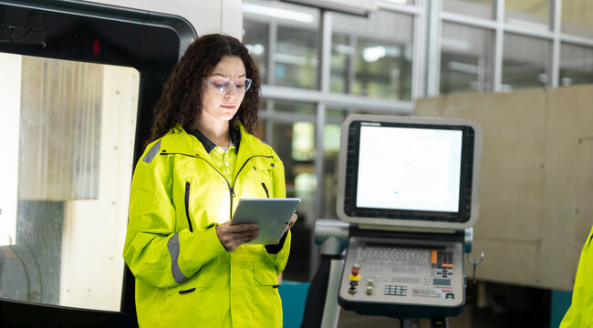 Women engineer wearing safety glasses uses a tablet beside a CNC machine control panel, showing precision manufacturing, industrial automation, quality inspection, smart factory technology.