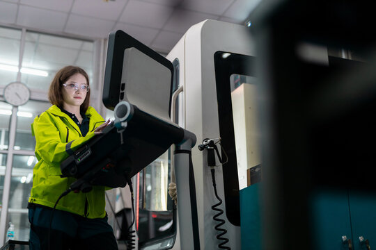 Female engineer in safety glasses operates a control panel on a CNC machine, showing industrial automation, precision manufacturing, machine setup, quality control, factory technology.