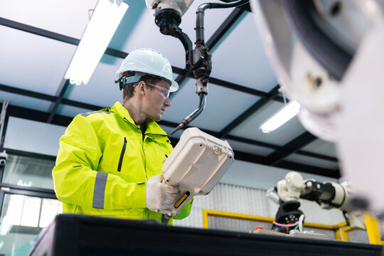 Man engineer in hardhat and safety glasses uses teach pendant to program industrial robot arm, showing automation, robotics control, factory safety, machine setup, manufacturing technology.