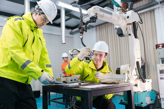 man engineers wearing hardhats and safety glasses inspect industrial robot arm in factory lab, showing automation maintenance, robotics repair, teamwork and manufacturing technology.