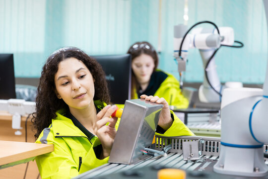 women engineer in safety jacket inspects a control box beside collaborative robot arm as teammate works, showing automation testing, robotics training, quality control, smart factory tech.