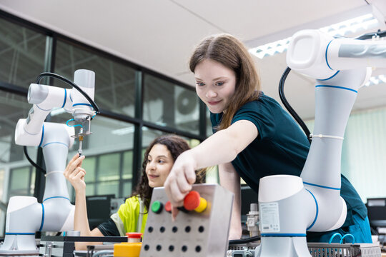 women engineer in safety jacket inspects a control box beside collaborative robot arm as teammate works, showing automation testing, robotics training, quality control, smart factory tech.