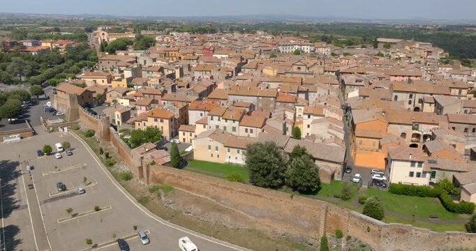 Panoramic drone view of the medieval walls of Tuscania built on a tuff cliff, in the province of Viterbo, in Lazio, Italy, with a large parking area, winding roads and the historic center above.