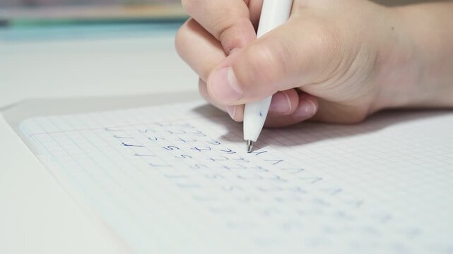 Child practices writing English alphabet letters with blue pen in graph paper notebook. Young learner carefully forms each character, focused on building early handwriting and literacy skills.