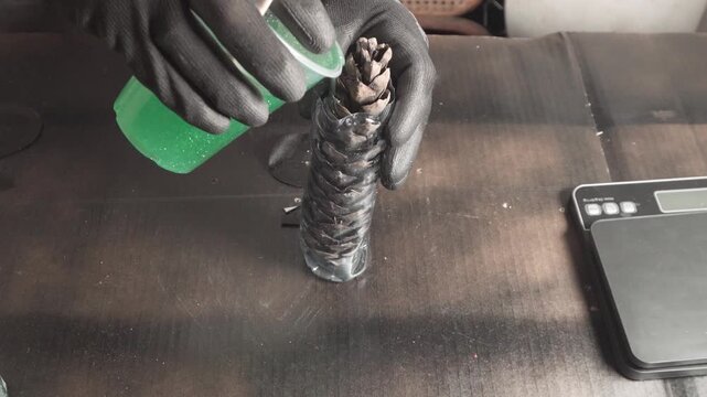 A craftsman in gloves carefully pours liquid green epoxy resin over a pine cone in a custom mold on a workbench, close-up shot.