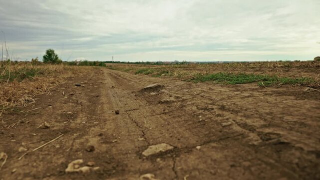 A low-angle shot of a dry field road and grass next to a plowed field under an overcast sky.