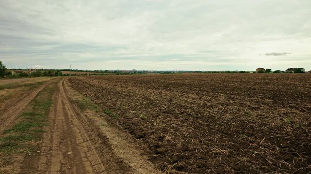 A wide shot shows a rural dirt road running alongside a plowed agricultural field under a cloudy overcast sky.