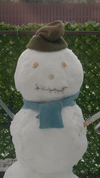 Vertical Screen: A snowman is built in the yard of an apartment building. It has a hat, arms made of tools, and a scarf. Snow covers the ground around it, showing a winter scene.