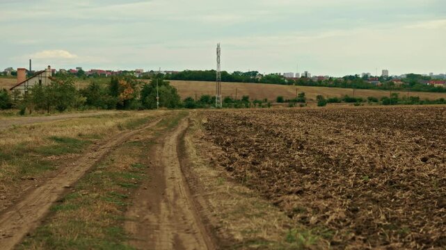 A wide shot shows a rural dirt road running alongside a plowed agricultural field under a cloudy overcast sky.