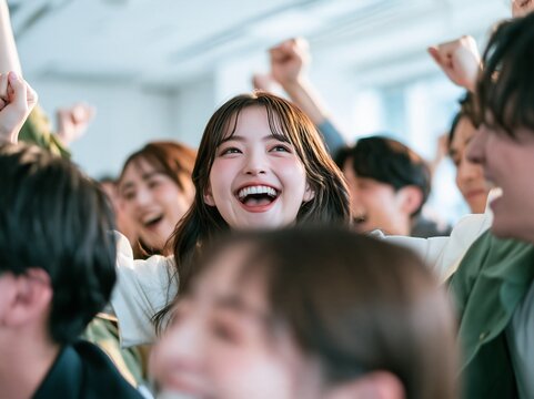 仲間たちと囲まれながら満面の笑みでガッツポーズを決める女性ビジネスパーソン。女性活躍推進の特集、社内報、若手育成セミナーに映える写真素材（文字入れ用余白・コピースペース付き）