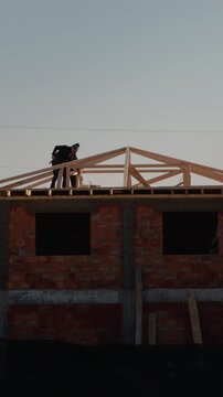 Vertical Screen: Workers are constructing the roof of a house. They are focused on placing wooden beams and ensuring the structure is stable. The work takes place in a residential area.