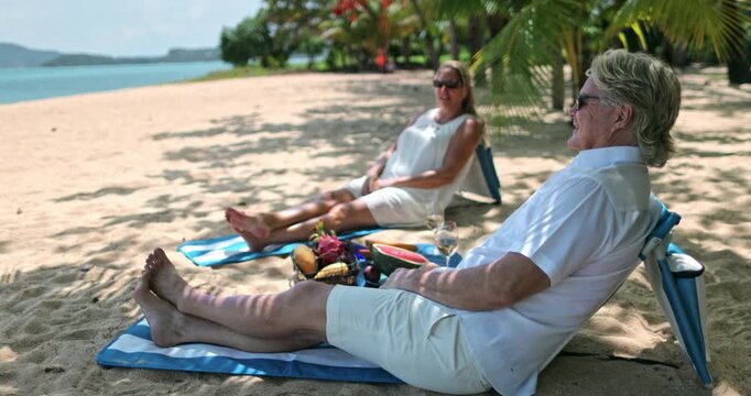 Senior couple enjoying a relaxing vacation on a tropical beach, sitting on towels in the sand with a picnic basket filled with exotic fruits and wine under palm tree shade. Koh Samui Island, Thailand