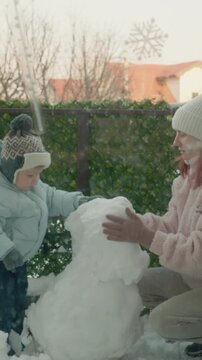 Vertical Screen: A mother and her young son work together to create a snowman in the yard of their apartment building on a snowy day. They enjoy their time playing in the snow.