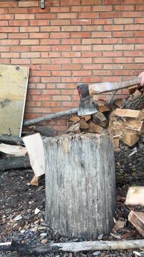 A person chopping firewood with an axe on a wooden chopping block outdoors. Close-up of splitting logs, with stacked firewood and a rustic brick wall in the background. Traditional wood preparation fo